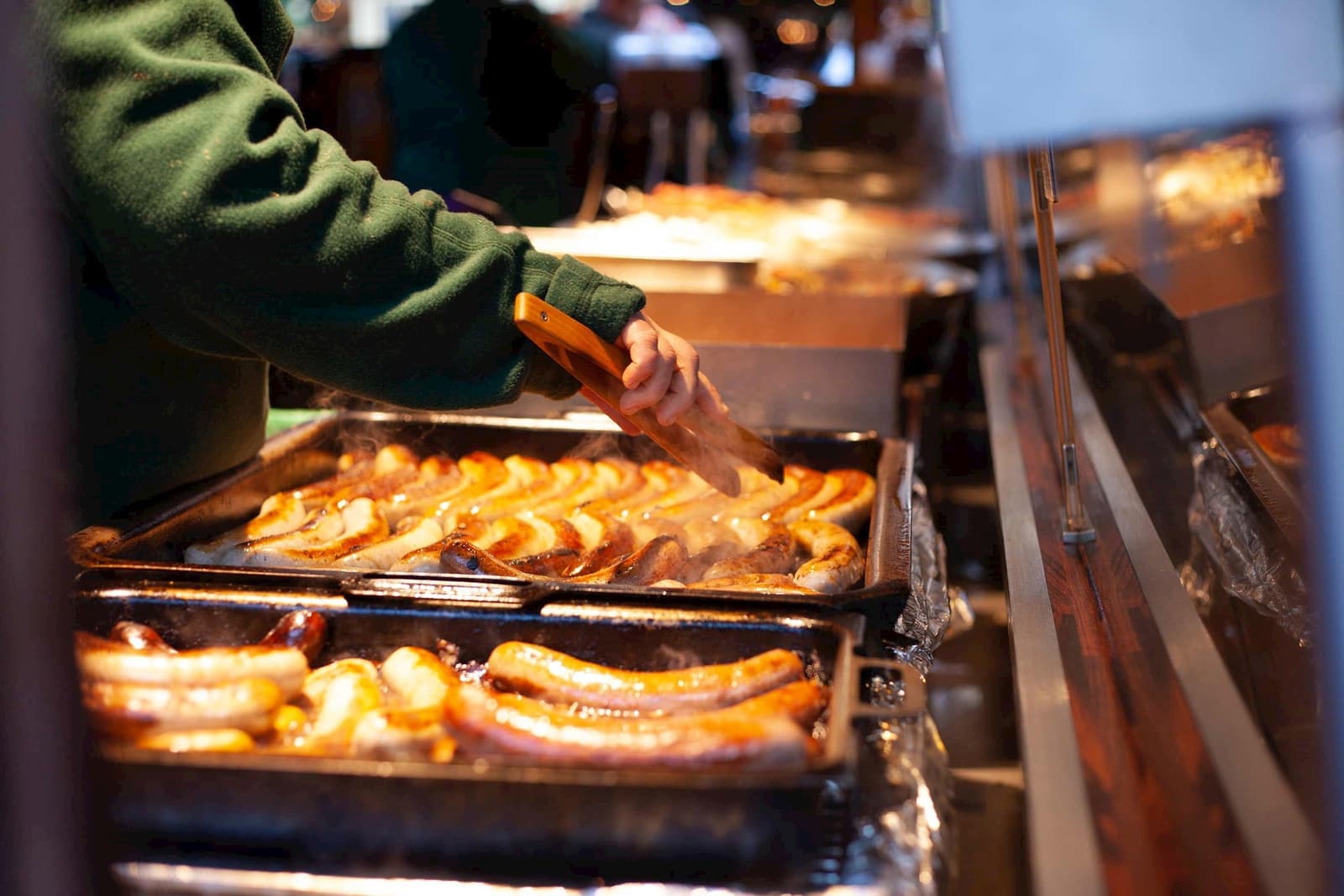 Hands using tongs to grill bratwurst sausages at food stall.
