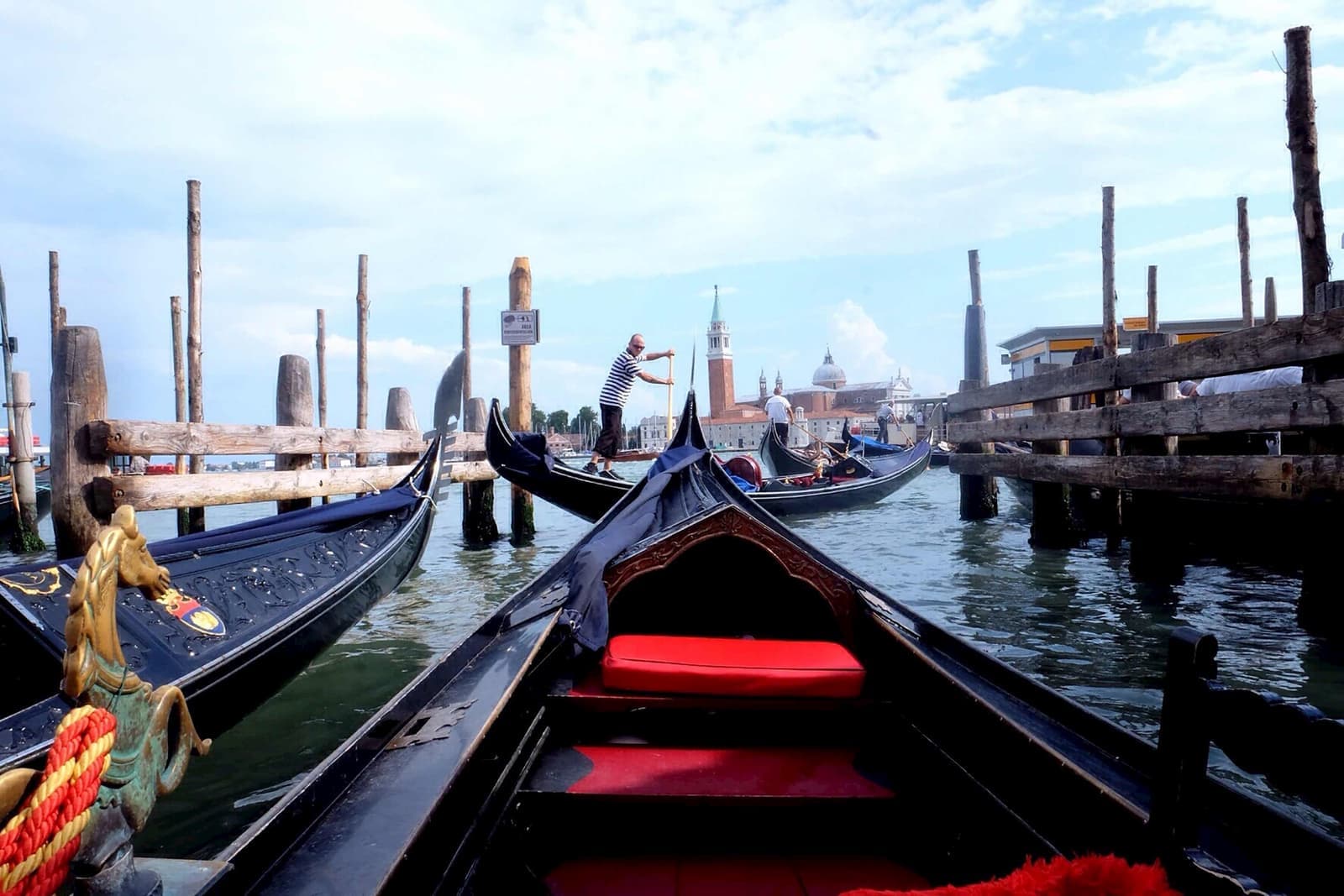 View from gondola dock in Venice with red cushioned seat, wooden poles and gondolier rowing in distance