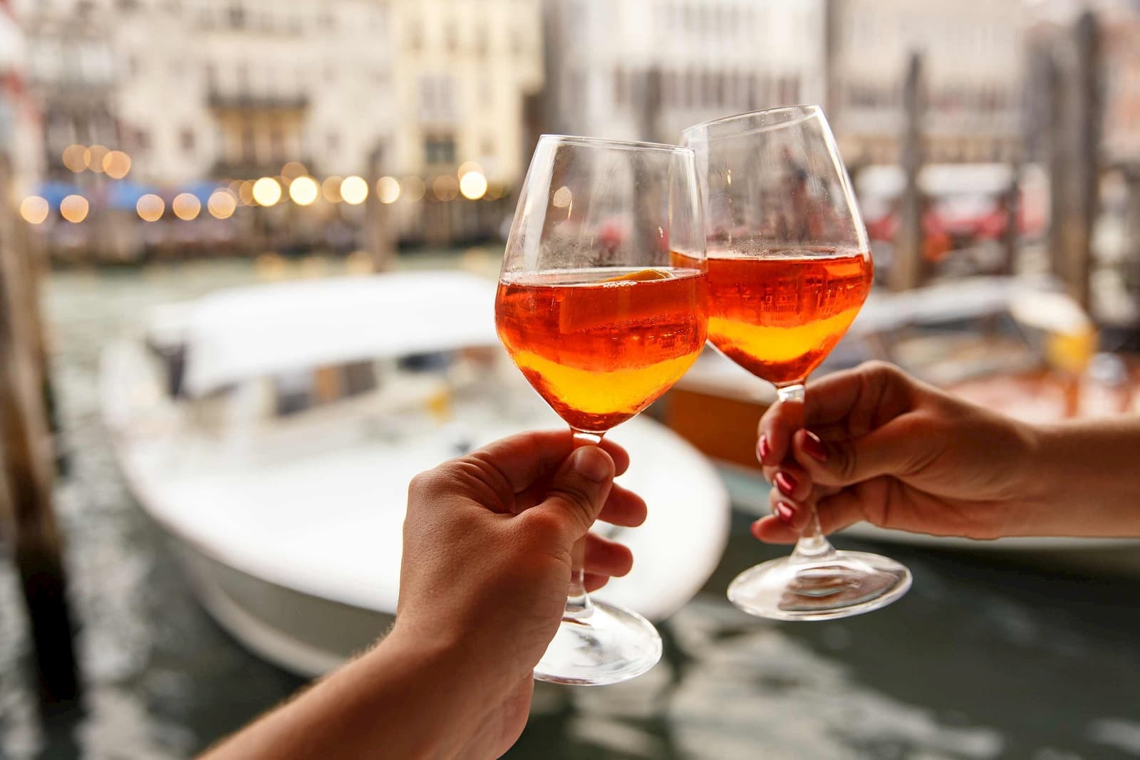 Two hands clinking glasses of Aperol Spritz with blurred boats in background