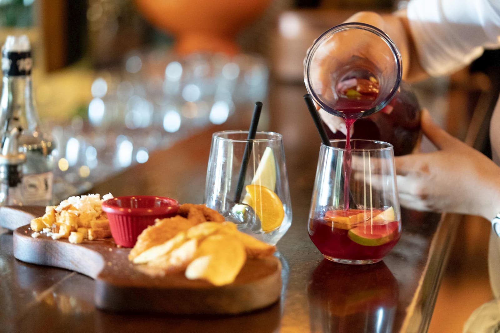 Person pouring red sangria with citrus slices into glass on bar counter beside tapas platter