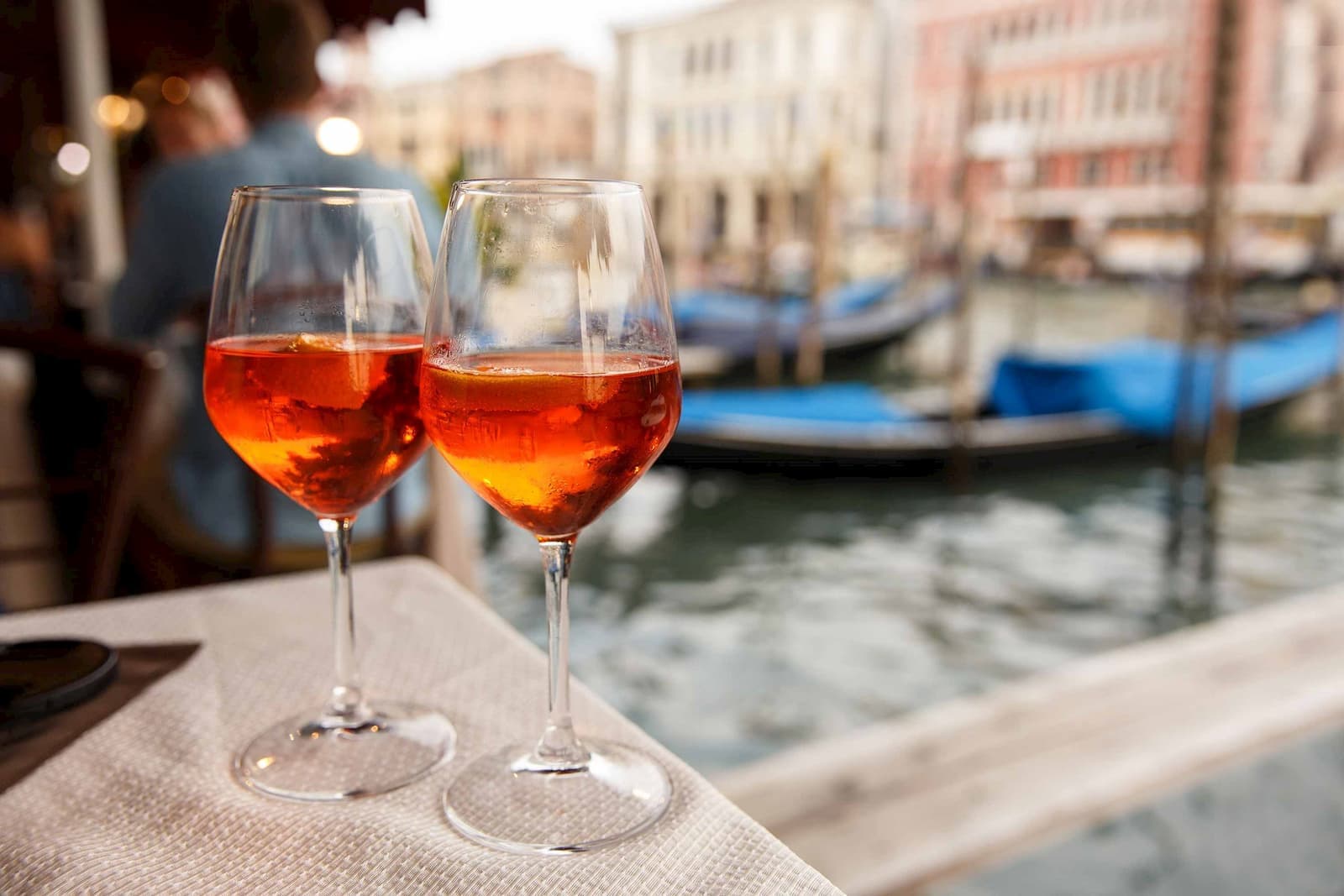 Two glasses of Aperol Spritz on outdoor table overlooking Venetian canal with gondolas