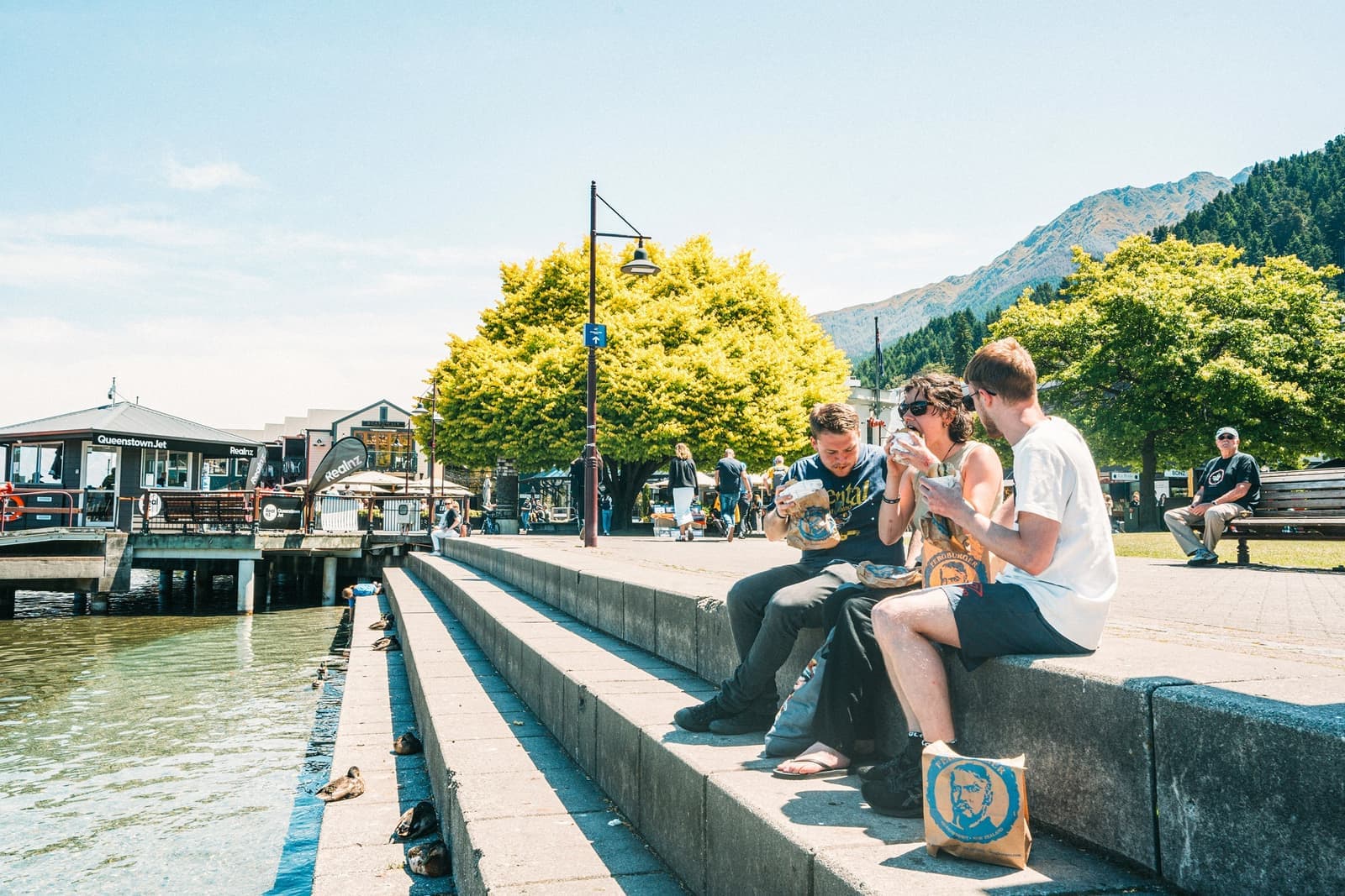 Three travellers enjoying burgers on steps next to waterfront, Queenstown, New Zealand