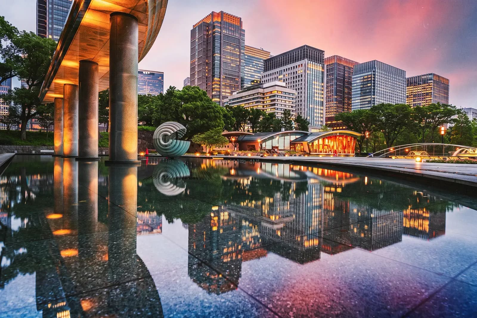 Tokyo skyline reflected in a plaza pool with office buildings and landscaped gardens.