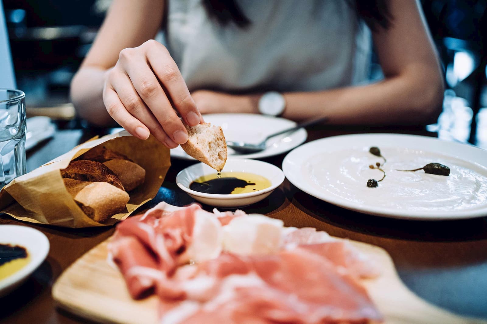 Hand dipping bread into olive oil beside cured meats and plates