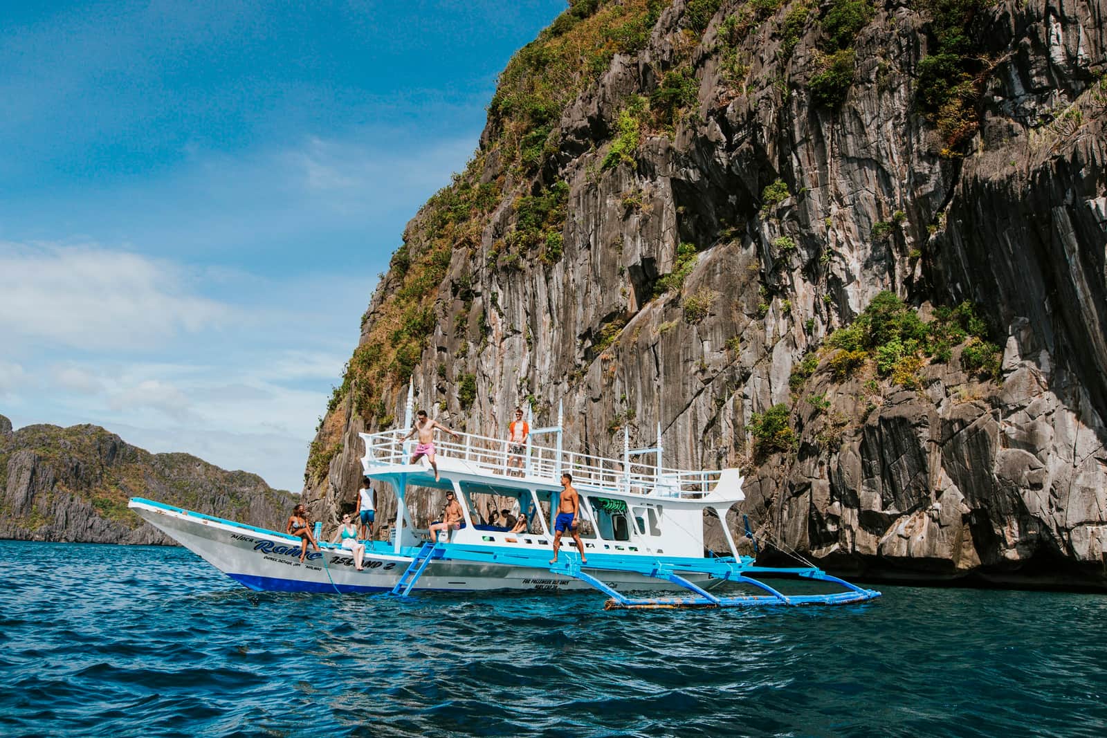 Travellers Relaxing and Jumping from White and Blue Boat Docked next to Cliff, El Nido, Philippines