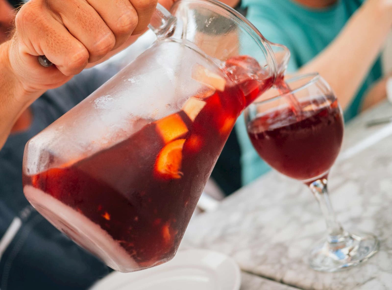 “Close-up of hand pouring sangria with fruit slices from glass pitcher into wine glass